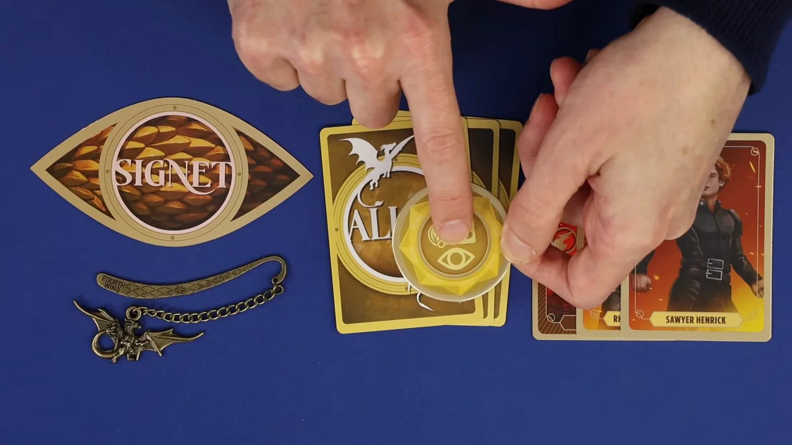 La carta SIGNET con forma de ojo en Rise of the Wingleader aparece junto a la ficha dorada marcada con el ojo que la desbloquea, con una carta de aliado y el marcapáginas de dragón de metal al lado.