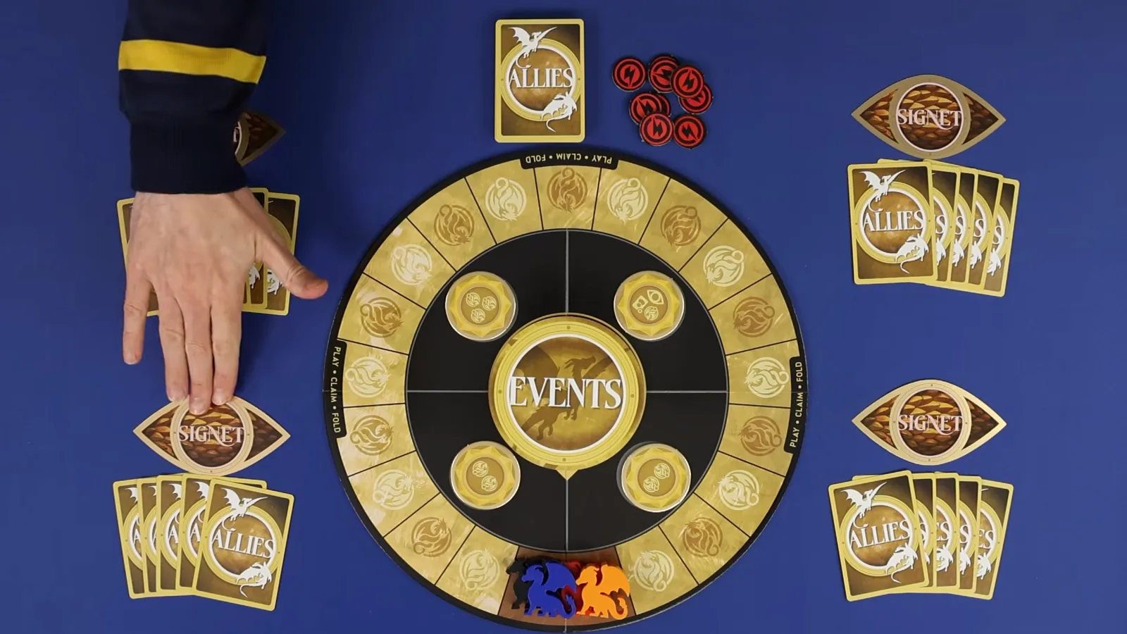 Durante la preparación de Rise of the Wingleader, un jugador coloca una carta SIGNET con forma de ojo boca abajo junto al tablero, con fichas rojas de herida en la parte alta de la mesa.